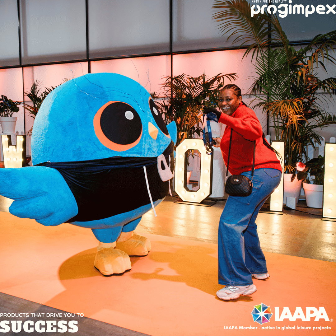 Person in a red jacket posing with a large blue cartoon bird mascot at an indoor event, with plants and illuminated letters in the background.